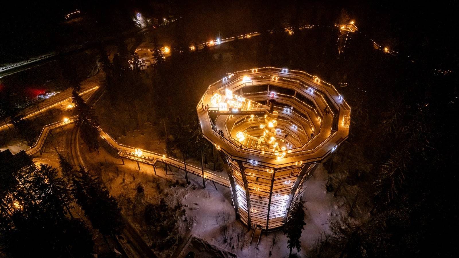 Osvetljena razgledna pot in stolp sredi zasneženega gozda ponoči.